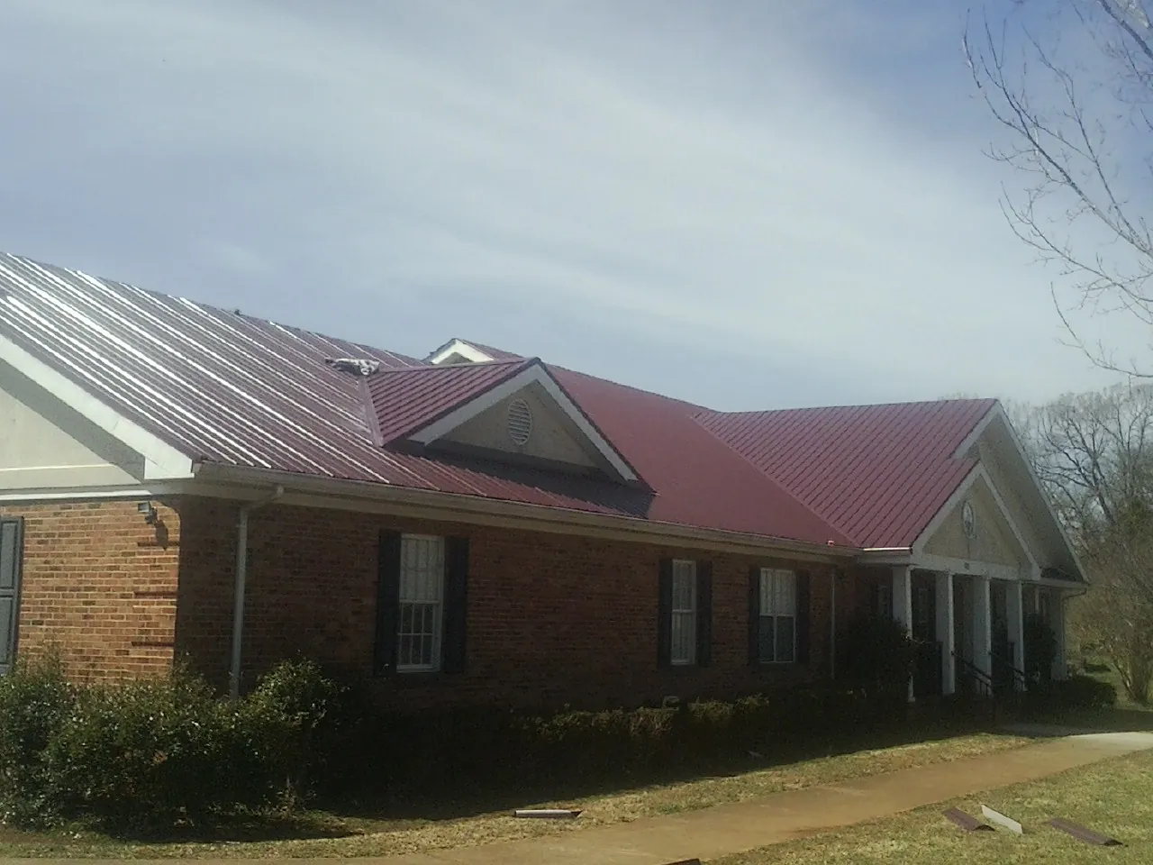 Red metal roof with gabled entries on a long brick building, from a Hail Damage Roof Repair job in Little Neck