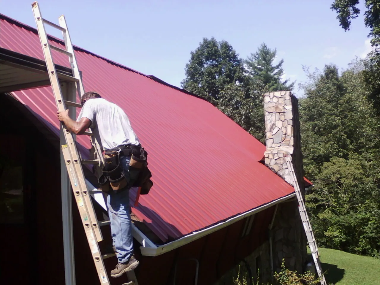 Roofer on a ladder beside a red metal roof and stone chimney — Soffit & Fascia Repair by Roofing Brothers in Little Neck