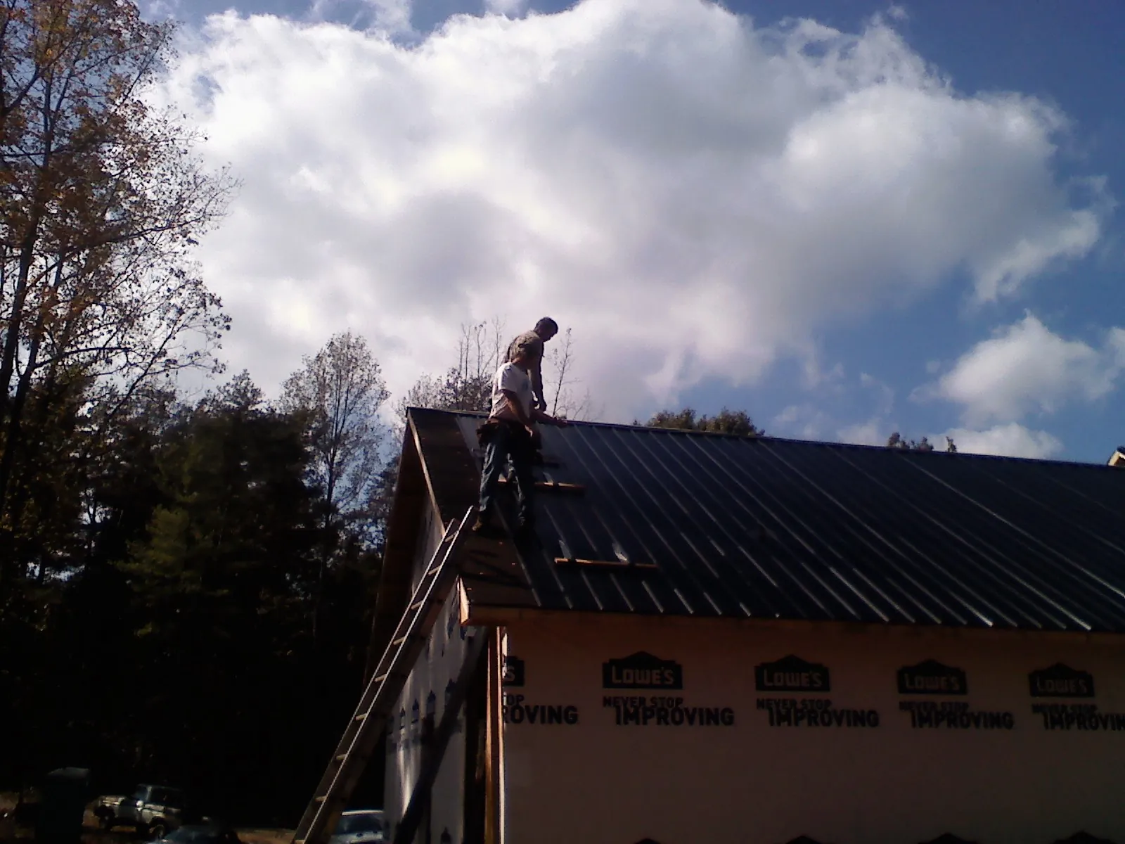Roofer fastening dark metal panels high against a cloudy sky, from a Roof Inspection job in Little Neck