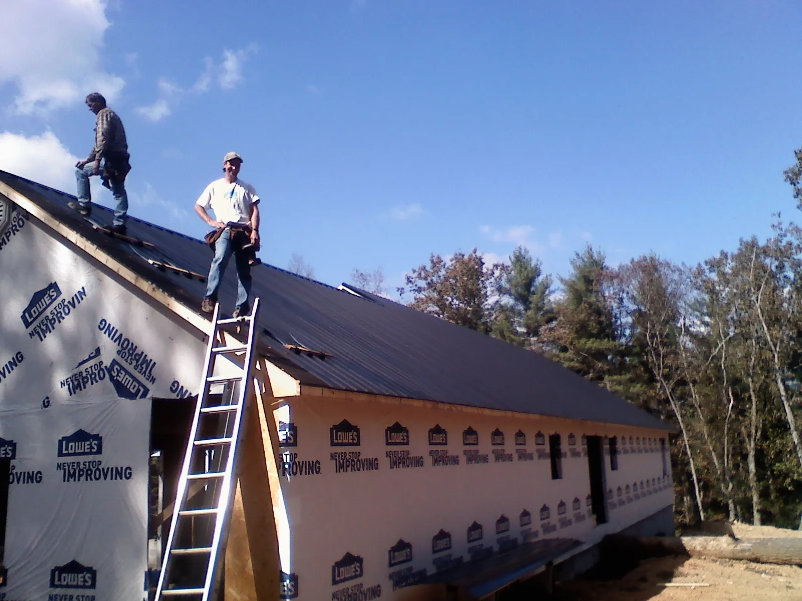 Two roofers standing on a long dark metal roof over a wrapped building in Little Neck